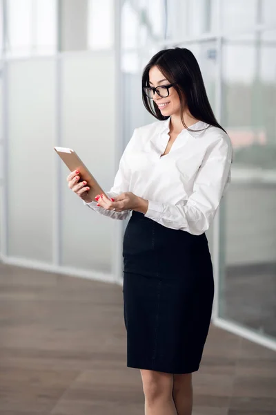 Atractiva joven mujer con gafas y leyendo su tableta de pantalla táctil mientras está de pie dentro del edificio comercial — Foto de Stock