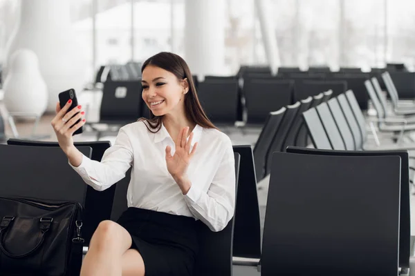 Empresaria viajera con teléfono inteligente, saludando con la mano en la cámara frontal durante la videollamada, esperando en el vestíbulo del aeropuerto. Mujer viajando al extranjero en viaje de negocios. Vuelo aéreo —  Fotos de Stock