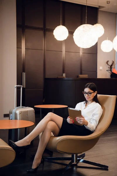Empresaria sentada en la sala de negocios del aeropuerto, esperando el vuelo. Mujer sonriente con gafas sentada en el sofá en la sala de espera del aeropuerto. —  Fotos de Stock