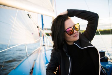 Güzel genç kadın yat kase üzerinde oturur ve pozlar. El ve gülümseme ile sunglusses giyer. Model yat gemide yelken.