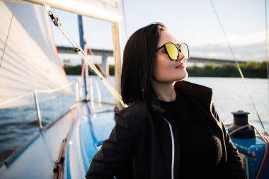 Güzel genç kadın yat kase üzerinde oturur ve pozlar. El ve gülümseme ile sunglusses giyer. Model yat gemide yelken.