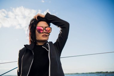 Güzel genç kadın yat kase üzerinde oturur ve pozlar. El ve gülümseme ile sunglusses giyer. Model yat gemide yelken.