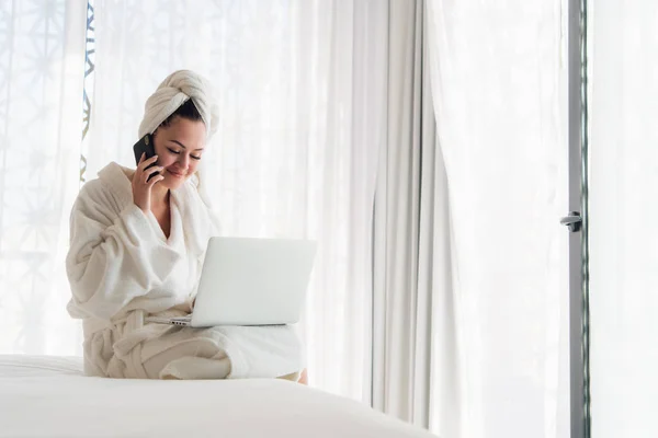 Mujer relajada con albornoz usando teléfono inteligente y portátil en la cama —  Fotos de Stock