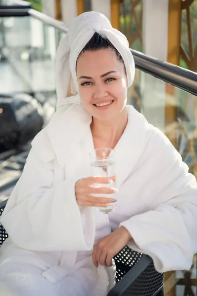 Mujer con vidrio. Jovencita alegre en un albornoz asistiendo al centro de spa y sonriendo mientras está sentada en la silla con un vaso de agua en la mano en la terraza —  Fotos de Stock