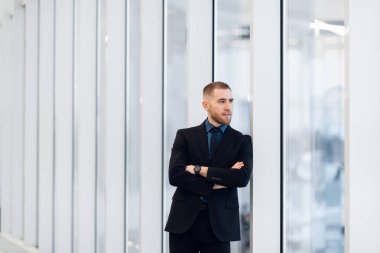 Orta yaşlı bir adam, kollarını kavuşturmuş, pencerenin kenarına yaslanmış kameraya bakıp düşünceli bir ifadeyle bakıyor.
