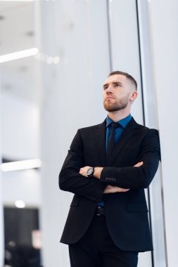 Orta yaşlı bir adam, kollarını kavuşturmuş, pencerenin kenarına yaslanmış kameraya bakıp düşünceli bir ifadeyle bakıyor.