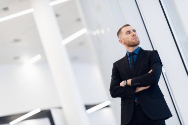 Orta yaşlı bir adam, kollarını kavuşturmuş, pencerenin kenarına yaslanmış kameraya bakıp düşünceli bir ifadeyle bakıyor.