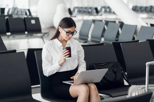 공항 의 출발 라운지에 앉아 노트북에서 작업 하는 커피를 가진 젊은 여자 — 스톡 사진