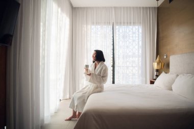 Bornoziçinde çekici genç kız bir fincan tutan, pencereye bakarak ve evde büyük pencereyakınında bir yatakta otururken gülümseyerek