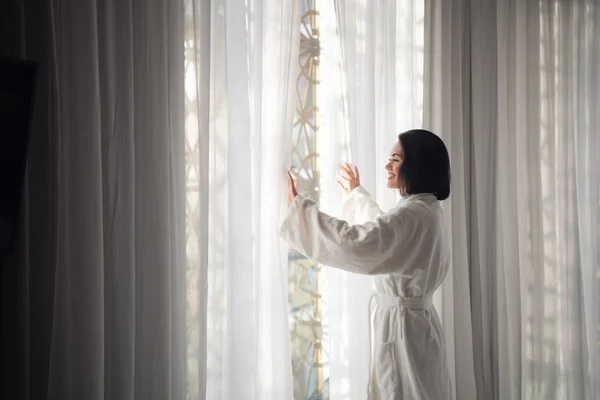 Mujer con albornoz blanco de pie por la mañana cerca de la ventana en la sala de luz —  Fotos de Stock