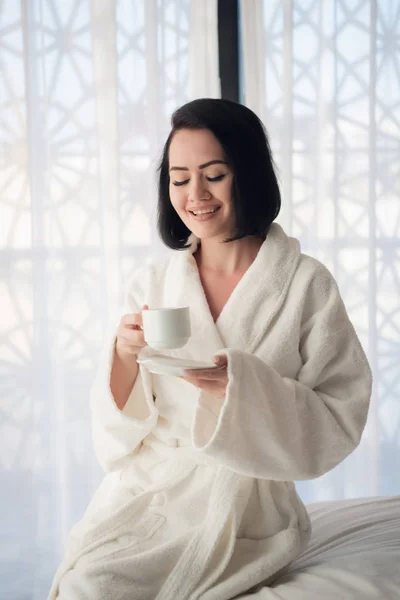 Una chica en un albornoz bebiendo café por la mañana en la cama en una sala de luz . —  Fotos de Stock