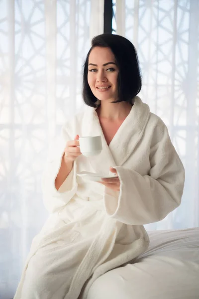 Muchacha joven atractiva en albornoz sosteniendo una taza, mirando a la ventana y sonriendo mientras se sienta en una cama cerca de la gran ventana en casa —  Fotos de Stock