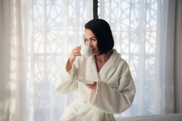 Una chica en un albornoz bebiendo café por la mañana en la cama en una sala de luz . —  Fotos de Stock