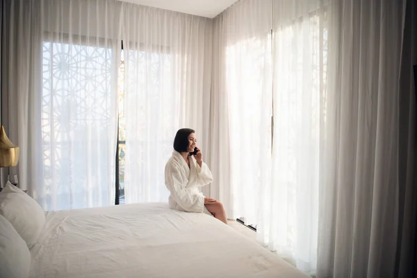 Mujer con albornoz blanco en la mañana cerca de la ventana en el cuarto de luz. Hablando por teléfono —  Fotos de Stock