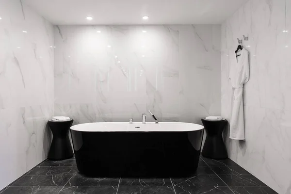 Freestanding black bathtub, stylish minimalist black and white loft style bathroom. Bath, washstand, walls with marble tiles.