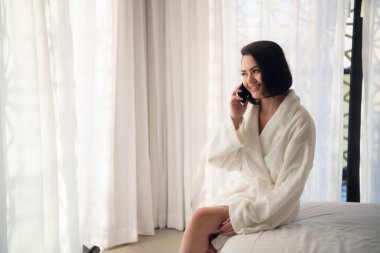 Sabahları beyaz bornoz giyen kadın ışık odasındaki pencerenin yanında. Telefonla konuşmak