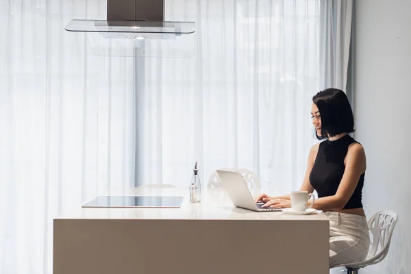 Hermosa mujer de negocios está utilizando un ordenador portátil, mirando a la cámara y sonriendo mientras se sienta en casa —  Fotos de Stock