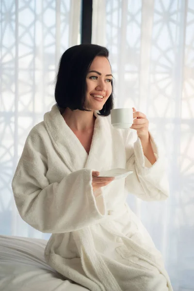 Muchacha joven atractiva en albornoz sosteniendo una taza, mirando a la ventana y sonriendo mientras se sienta en una cama cerca de la gran ventana en casa —  Fotos de Stock