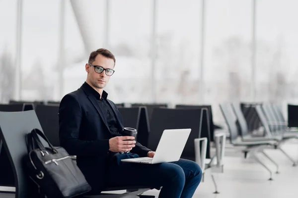 Joven hombre de negocios guapo trabajando en una computadora portátil y bebiendo un café durante una expectativa de un vuelo —  Fotos de Stock