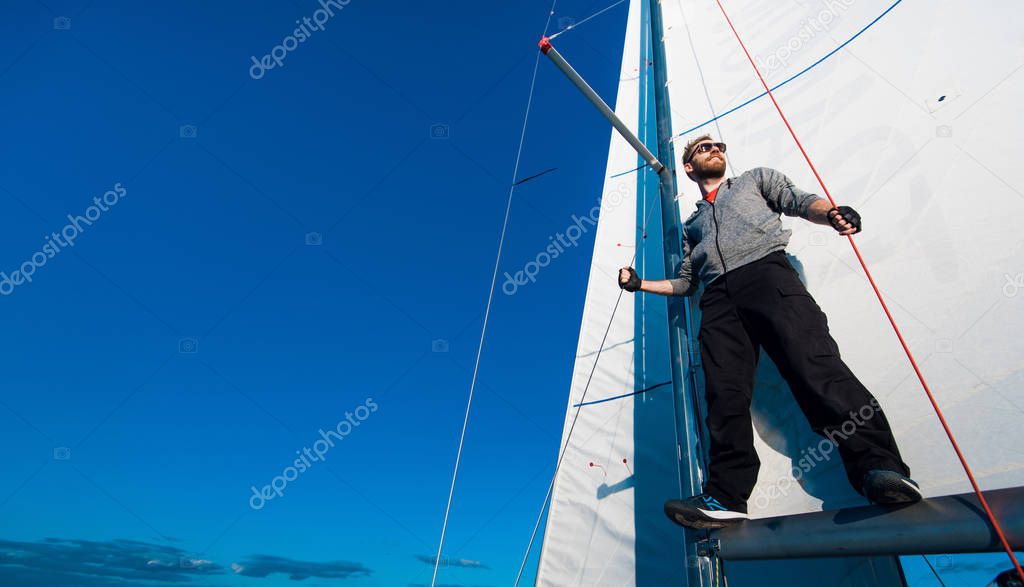 Positive skilled young male sailor with beard wearing sunglasses adjusting sail on boat and examining it while fixing sail on boom.