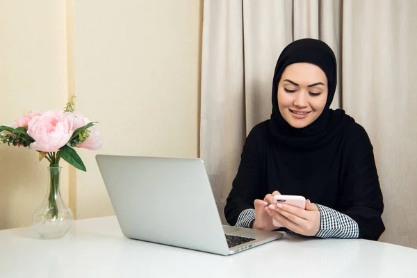 Mujer musulmana hablando por teléfono mientras trabaja con computadora portátil . — Foto de Stock