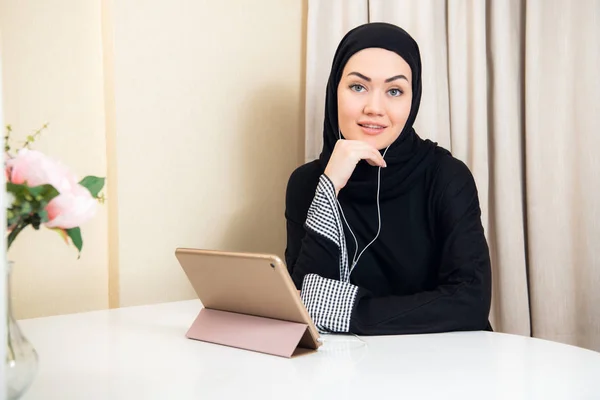 Mujer musulmana sentada en casa con tableta, con auriculares. Descanse en casa, preparándose para ver una película o escuchar una música . — Foto de Stock