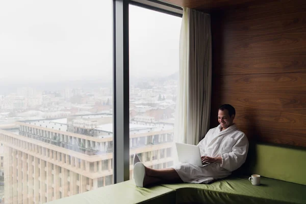 El joven está trabajando en un portátil en su cama sobre un fondo de una ventana panorámica con vistas a los rascacielos. Freelancer, trabajo remoto, trabajo desde casa . —  Fotos de Stock