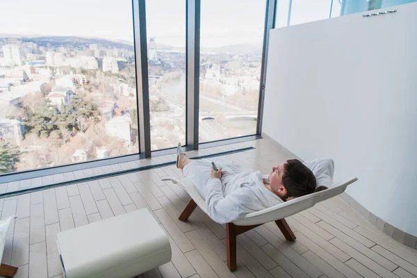 Joven descansando en una moderna tumbona frente a una gran ventana con vista a la ciudad en un moderno spa —  Fotos de Stock