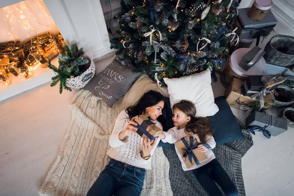 Linda niña y su madre jugando bajo el árbol de Navidad con regalos, acostada en el suelo de madera en la habitación. El uso de suéter de punto de moda, jeans. Vista superior. —  Fotos de Stock