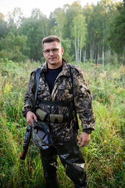 Avcı açık havada av tüfeğiyle gözlük ve kamuflaj takıyor.