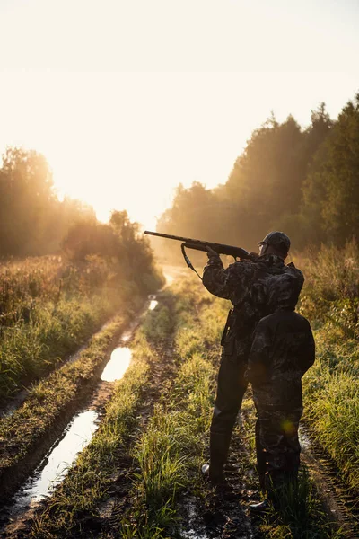 步枪猎人和他的儿子轮廓在美丽的日落。 日出时分，猎人带着男孩和来复枪在森林里. — 图库照片