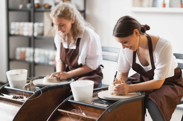 Overview of two young potters by pottery wheels while preparing ceramic pots from raw clay.