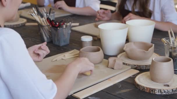 In the clay workshop. Close up of female hands during the working process in the clay workshop. Female potter masters rolling up the clay on table with ceramic products — Stock Video