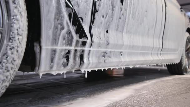 Biltvätt. Bilen trösklar är täckt med vitt flödande skum vid en biltvätt. Närbild med en suddig bakgrund. — Stockvideo