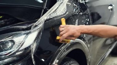 Siyah bir arabanın üzerine boya koruma filmi montajı. Master, film kenarlarını düzgün eğmek için cam sileceği aracı kullanarak poliüretan koruyucu film uyguluyor