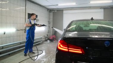 Adam araba tamirhanesinde araba yıkamak için yüksek basınçlı çamaşır makinesinden su püskürtüyor..