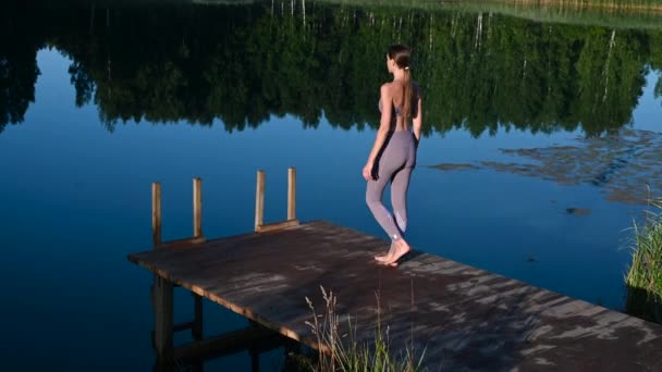 Young woman walks on wooden pier above forest lake scenery, folds her arms in a namaste gesture. Woman arms outstretched in nature. — Stock Video