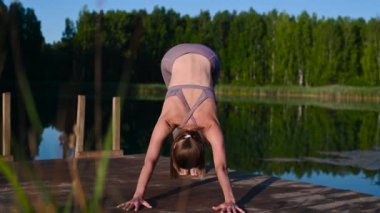 Gölün kenarında yoga yapan genç bir kadın ahşap platformda hareket ediyor. Doğada egzersiz yapan güzel bir genç kadın, sağlıklı bir yaşam tarzı genç insanlar pozitif titreşimler alıyor..