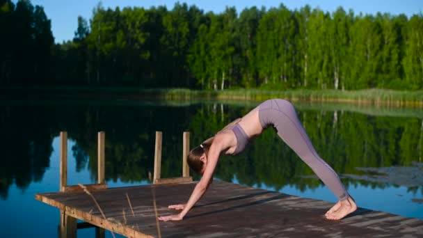 Mujer joven junto al lago practicando yoga se mueve sobre plataforma de madera. Mujer bastante joven haciendo ejercicio en la naturaleza, estilo de vida saludable jóvenes vibraciones positivas. — Vídeo de stock