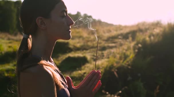 スポーツ服を着たヨガの女性が瞑想し、自然の中で日の出を呼吸します。手に芳香族スティックから煙. — ストック動画