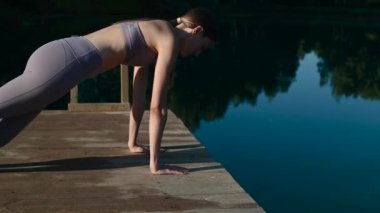 Genç çekici kız gün batımında göl kenarında yoga yapıyor. Suyun kenarında lotus pozisyonunda oturuyor..