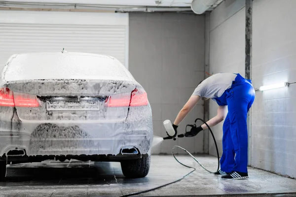 Coche con luces de fondo encendidas de pie en el servicio de lavado de coches. Trabajador cubrir el coche con espuma con pulverizador de alta presión. — Foto de Stock