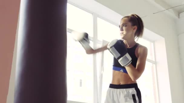 En tjej i sportkläder är engagerad i boxning och tränar ett slag med handen på en boxningssäck — Stockvideo