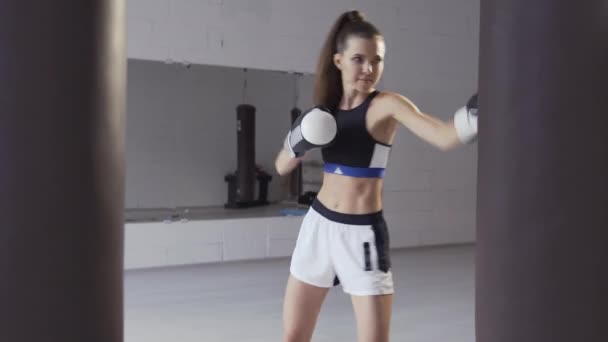 Una bonita chica kickboxer se está preparando para las competiciones en la sala de boxeo, practicando la técnica de golpes en un saco de boxeo — Vídeos de Stock