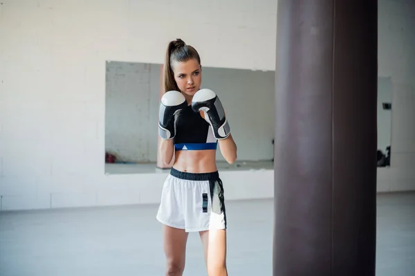 La chica se está preparando para una competición de boxeo y entrena golpes en un saco de boxeo en un amplio gimnasio — Foto de Stock