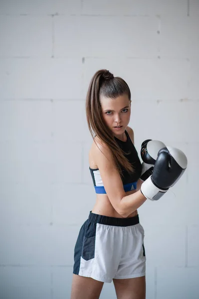 Una hermosa atleta con guantes de boxeo y uniforme deportivo se levanta contra una pared de ladrillo blanco — Foto de Stock
