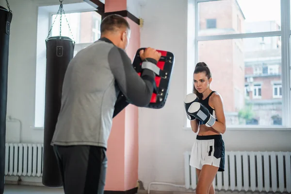 Riscaldamento prima dell'allenamento. Una ragazza in una posizione di boxe con i guanti si prepara a colpire le zampe nelle mani dell'allenatore. — Foto Stock