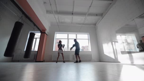 손에 권투 발을 쥐고 있는 한 남자는 가벼운 체육관에서 초보 소녀를 때리는 기술을 가르친다 — 비디오