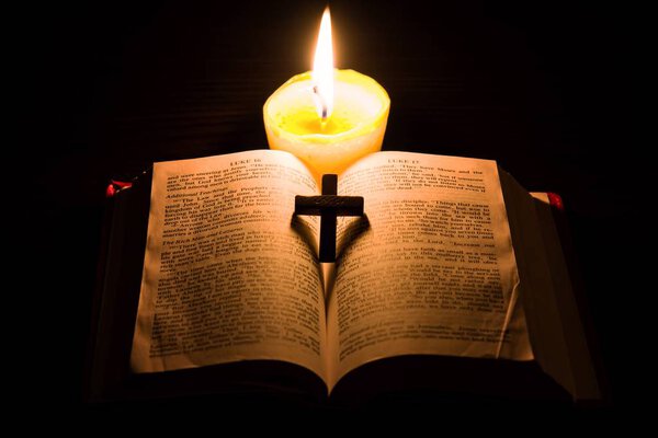 Burning candle and Bible . The psalms . Holy book . told in the Bible's New Testament , on a black background . Christian cross .