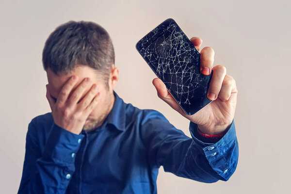 The guy is holding a black smartphone with a broken display. Broken screen of modern frameless phone. guy crying with his eyes closed and hands in his face.